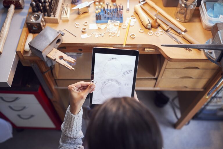 overhead-view-of-female-jeweller-comparing-ring-with-drawn-design-on-digital-tablet-in-studio